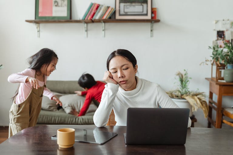 A tired homeowners mother working remotely on her laptop while children play in the background.