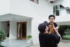 A father playfully lifting his child outside a modern home on a sunny day.