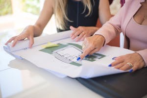 Two women reviewing detailed architectural blueprints in a professional setting. an architect consulting with homeowners, pointing to a blueprint with sustainable design elements highlighted.